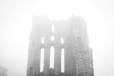 Uzun, dar pencereleri ve kemerleri olan antik bir taş binanın kalıntılarını içeren sisli bir sahne. Yapı kısmen çökmüş görünüyor ve yoğun sis arkaplanı gizliyor. Bu da gizemli ve ürkütücü bir atmosfer yaratıyor..