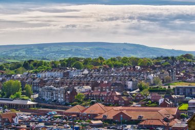 Parlak bulutlu gökyüzünün altında yemyeşil tepeler ve ağaçlarla çevrili sıra sıra evler ve binalarla dolu küçük bir kasabanın manzarası..