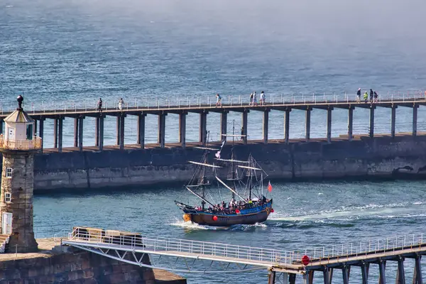 stock image A historic sailing ship with multiple masts and sails is navigating through a harbor with a pier and lighthouse in the background. People are walking on the pier, and the water is calm.