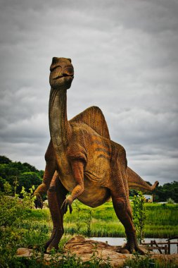 Açık hava parkında duran büyük bir dinozor heykeli. Arka planda yeşil ve bulutlu gökyüzü var..