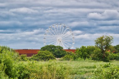 Arkasında bulutlu bir gökyüzü olan yeşil ağaçların ve çalıların üzerinde yükselen büyük bir dönme dolap..