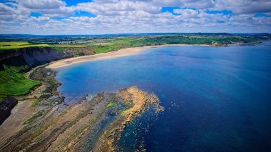Açık mavi su, kayalık kıyılar ve parçalı bulutlu bir gökyüzünün altındaki yeşil alanlar manzaralı bir kıyı şeridi..