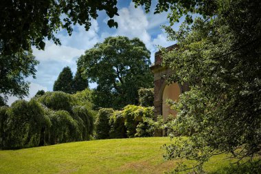 Bereketli yeşillik, ağaçlar ve kısmen görünen tarihi tuğla kemerli bir park sahnesi. Gökyüzü parçalı bulutlu, sakin atmosfere ekleniyor..