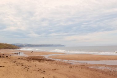 Sahilde birkaç kişinin yürüdüğü sakin bir sahil sahnesi. Gökyüzü bulutlu ve dalgalar kumlu sahilde hafifçe yuvarlanıyor. Uzakta kayalıklar ve puslu bir ufuk var..