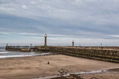 Denize uzanan iki deniz feneri olan uzun bir rıhtım manzarası. Ön plandaki kumlu plaj çoğunlukla boş, suyun yanında birkaç kişi görünüyor. Gökyüzü bulutlu, sakin ve sakin bir atmosfer yaratıyor..