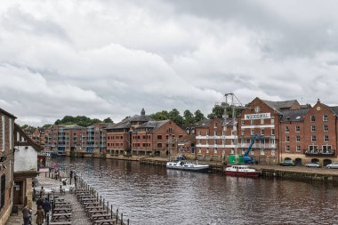 Nehir kenarında tuğla binalar, su boyunca yanaşmış tekneler ve bulutlu bir gökyüzü..