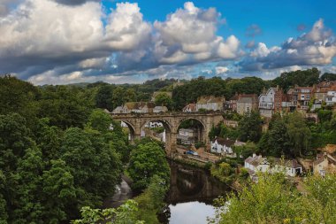 Nehir üzerindeki tarihi bir taş köprünün manzarası. Etrafı yemyeşil ve bulutlu bir gökyüzünün altındaki antika evlerle çevrili..
