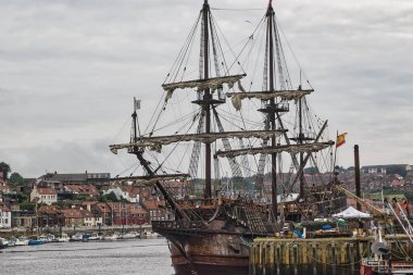 Tarihi bir yelkenli gemi arka planda bir kasabayla birlikte limana yanaştı. Geminin birden fazla direği ve donanımı var ve liman binalar ve teknelerle çevrili..