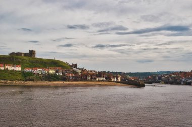 Bir tepenin üzerindeki tarihi binaların, kumlu bir plajın ve bulutlu bir gökyüzünün altında sakin bir su kütlesinin manzarası..