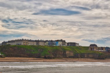 Kumlu bir sahile ve okyanusa tepeden bakan bir dizi binası olan bir sahil kasabası. Gökyüzü bulutlu ve birkaç kişi sahilde yürüyor..
