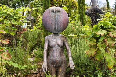 Büyük yuvarlak bir kafası ve minimalist bir tasarımı olan, yemyeşil ve bahçedeki bitkilerle çevrili, eşsiz bir insansı heykel..