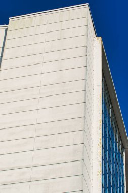 Modern bir binanın arka planında açık mavi gökyüzü olan beton bir cepheyi gösteren yakın plan görüntüsü. Bina, cam pencerelerden yansıyan pürüzsüz ve dokulu yüzeylerden oluşan bir karışıma sahip..