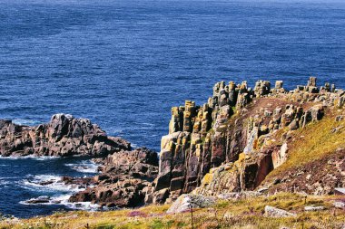 Derin mavi bir okyanusa bakan yosun ve liken kaplı engebeli uçurumlar ve kayalık oluşumların yer aldığı manzaralı bir kıyı manzarası. Manzara, kayalık araziye karşı canlı bir kontrast oluşturan kır çiçekleriyle dolu..
