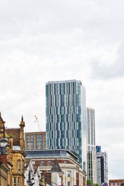 Camdan bir cephesi olan modern bir gökdelen eski binaların arasında kentsel bir ortamda duruyor. Gökyüzü bulutlu, şehir manzarasına dramatik bir zemin ekliyor..