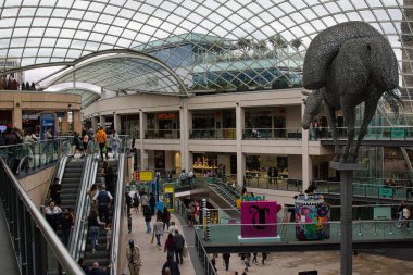 Camdan çatılı hareketli bir alışveriş merkezi, yürüyen merdivenler ve büyük bir ayı heykeli. Alışveriş yapanlar çeşitli mağazaları ve vitrinleri gezerken görülürler..