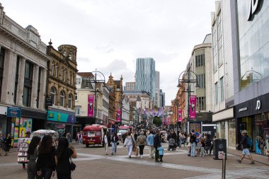 Yayalar, dükkanlar ve renkli afişlerle dolu hareketli bir şehir caddesi. Mimaride, arka planında belirgin bir gökdelen bulunan modern ve tarihi binaların bir karışımı yer alıyor. Atmosfer canlı ve kentsel..