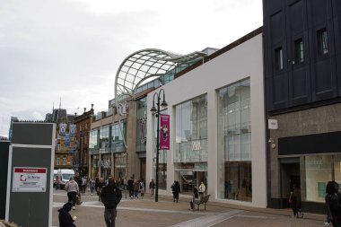 Modern mimarinin, büyük cam mağazaların ve yürüyen yayaların bulunduğu hareketli bir alışveriş caddesi. Sahnede, perakende mağazaları ve çağdaş bir tasarımla birlikte pankartlar ve pankartlar yer alıyor..