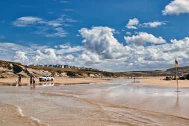 Sahilde yürüyen insanlarla manzaralı bir sahil manzarası. Gökyüzü kabarık bulutlarla dolu ve güneş ışıl ışıl parlıyor. Arka planda bir tepede binalar var. Bir cankurtaran aracı kumsalda park halinde ve güvenlik bayrağı görünüyor..