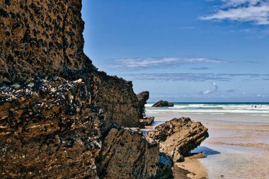 Engebeli kaya oluşumları ve nazik dalgaların olduğu manzaralı bir sahil manzarası. Kayalar, kumlu sahil ve açık mavi gökyüzü ile çelişen desenli ve koyu renklidir. Okyanus sakin, kumsalın tadını çıkaran birkaç uzak figür var..