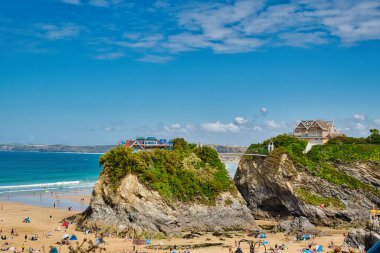 Kayalık kayalıkların, kumlu sahillerin ve güneşin tadını çıkaran insanların olduğu manzaralı bir sahil manzarası. Gökyüzü parlak mavi, dağınık bulutlar ve okyanusa bakan kayalıklara tünemiş evler var..
