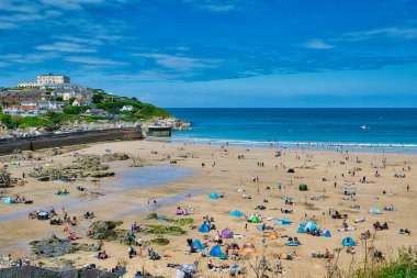 Sayısız insanın kumlu bir sahilde güneşin tadını çıkardığı canlı bir sahil sahnesi. Renkli sahil çadırları ve şemsiyeler manzarayı işaretlerken, hafif dalgalar kıyıda tur atıyor. Arka planda, beyaz binaları ve yeşillikleri olan bir kıyı kasabası görülebiliyor.