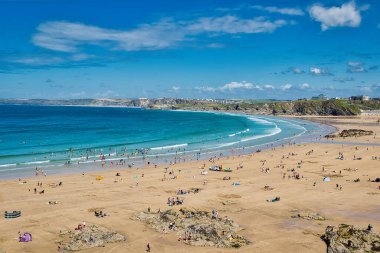 Altın kumu ve berrak mavi suları olan manzaralı bir sahil manzarası. İnsanlar yüzmek ve güneşlenmek gibi çeşitli aktivitelerden hoşlanıyorlar. Sahil şeridinde hafif dalgalar ve kayalık oluşumlar vardır, parlak mavi gökyüzünün altında birkaç bulut vardır..
