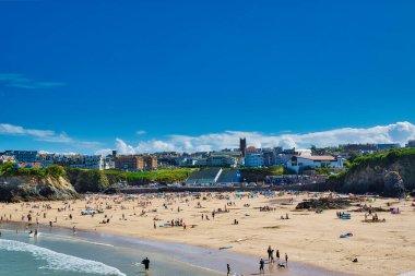 Güneşin tadını çıkaran insanlarla dolu kumlu bir sahil sahnesi. Arka planda binaları ve açık mavi gökyüzü olan bir kasaba yer alıyor. Bazı sahile gidenler güneşlenirken, diğerleri yürüyor ya da oynuyor..