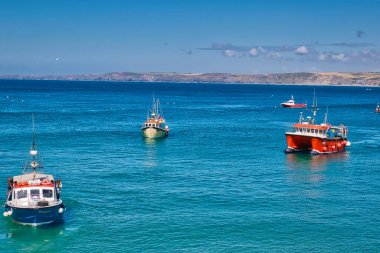 Birkaç balıkçı teknesiyle sakin bir denizin manzarası. Tekneler kırmızı ve beyaz dahil olmak üzere çeşitli renklerdedir ve berrak mavi suyla çevrilidir. Arka planda yuvarlanan tepeler ve birkaç bulutla birlikte açık mavi bir gökyüzü var.