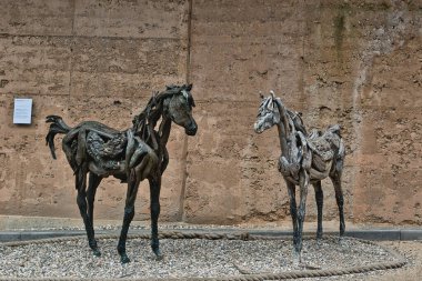 Çarpık ve dokulu malzemelerden yapılmış iki sanatsal at heykeli, dokulu bir duvara dayanıyor. Atlar birbirlerine dönük bir şekilde konumlandırılmışlar, şekillerinde karmaşık detaylar sergiliyorlar..