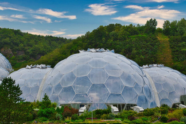 A view of large, geodesic domes made of glass and metal, surrounded by lush greenery and hills under a blue sky with clouds. The domes are part of a botanical garden or ecological project.