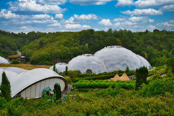 A scenic view of a botanical garden featuring large geodesic domes surrounded by lush greenery and trees under a partly cloudy sky.