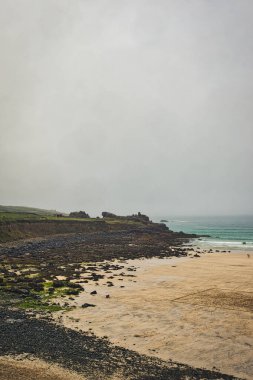 Sakin bir sahil sahnesi kayalık bir kıyı şeridi, yumuşak dalgalar ve bulutlu bir gökyüzü. Kum açık renklidir ve kayaların arasında yeşil yosun parçaları vardır. Uzakta, plajın tadını çıkaran birkaç kişi görülebilir..
