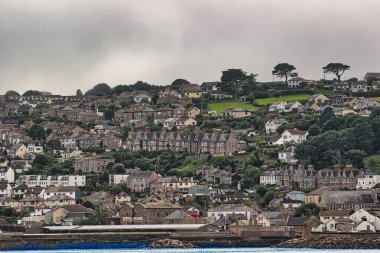 Kalabalık evleri olan bir dağ kenarı manzarası mimari tarzların bir karışımını sergiliyor. Çevrede yeşil tepeler ve bulutlu bir gökyüzü var, sakin bir atmosfer yaratıyor..
