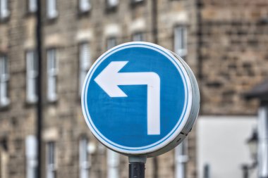 A blue circular traffic sign indicating a left turn, with a white arrow pointing left. The background features blurred buildings, suggesting an urban setting. clipart