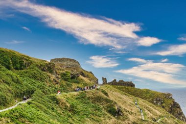 Bereketli yeşilliği ve dolambaçlı yolu olan bir tepe manzarası. İnsanlar eski harabelerin yakınındaki yolda yürüyorlar. Parlak mavi bir gökyüzünün altında, dağınık bulutlarla..