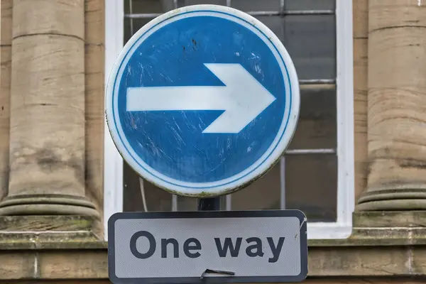 stock image A circular blue road sign with a white right arrow, indicating one-way traffic. Below it, a rectangular sign reads 'One way'. The background features a historic building with stone columns.