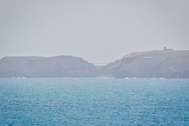Uçurumların ve ufuktaki uzak bir yapının yer aldığı puslu bir kıyı manzarası. Okyanus, yumuşak dalgalarla sakin, sakin bir atmosfer yaratıyor..