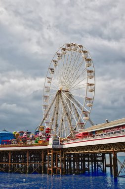 Rıhtımda büyük bir dönme dolap duruyor, renkli eğlence gezileri ve eğlencelerle çevrili. Gökyüzü bulutlu, aşağıdaki sakin suyu yansıtıyor..