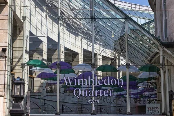 Tavandan sarkan renkli şemsiyelerin yer aldığı modern bir cam yapı ve 'Wimbledon Quarter SW19' metni belirgin bir şekilde sergileniyor. Sahne hareketli bir alışveriş alanı yakalıyor..