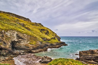 Engebeli uçurumlar, yemyeşil tepeler ve sakin bir deniz manzaralı bir sahil manzarası. Gökyüzü parçalı bulutlu, sakin bir atmosfer yaratıyor..
