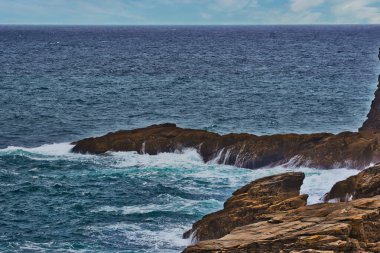 Bulutlu bir gökyüzünün altında kayalara çarpan kayalık kıyı şeridi manzarası. Okyanus koyu mavi, engebeli kıyı şeridiyle çarpıcı bir tezat oluşturuyor..