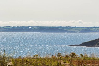 Işıldayan mavi suların, arka planda yuvarlanan yeşil tepelerin ve berrak bir gökyüzünün olduğu sakin bir kıyı manzarası. Sahne, doğanın güzelliğini huzurlu bir atmosferle yakalıyor..