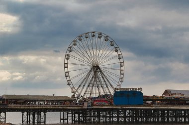 Büyük bir dönme dolap bir rıhtımda duruyor, eğlence gezileri ve oyunlarla çevrili. Gökyüzü bulutlu, karamsar bir atmosfer yaratıyor. Sahne bir nostalji ve eğlence karışımı yakalıyor, tipik deniz kenarı eğlenceleri..