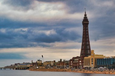 Blackpool 'un ikonik Blackpool Kulesi' nin dramatik bir gökyüzüne karşı manzarası. Plaj ön planda görülebiliyor. İnsanlar deniz kenarında eğleniyor. Gezinti alanı binalarla dolu..