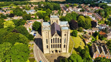 Yeşillik ve tuhaf bir köyle çevrili büyük tarihi bir katedralin havadan görünüşü. Katedralde karmaşık mimari ve yüksek kuleler yer alırken, köyde geleneksel evler ve ağaçlarla kaplı caddeler sergileniyor..