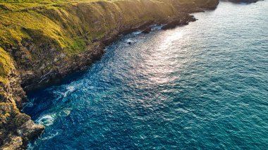 Uçurumları ve berrak mavi okyanus suyu olan engebeli bir sahil manzarası. Güneş ışığı sudan yansıyarak parıldayan bir etki yaratıyor. Manzarada kayalıklarda yeşil çimenler ve kıyı boyunca kayalık oluşumlar yer alıyor..