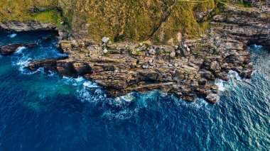 Açık mavi suları ve dalgaları kayalara çarpan kayalık kıyı şeridinin hava manzarası. Manzarada yeşil tepeler ve dağınık kayalar var, dramatik bir doğal manzara yaratıyor..