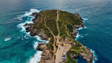 Aerial view of a rugged coastal island with green hills, winding paths, and rocky shores. The surrounding ocean features waves crashing against the rocks, creating a picturesque landscape. A few vehicles are parked near the base of the island. clipart