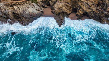 Kayalık kıyı şeridine çarpan okyanus dalgalarının hava görüntüsü, canlı turkuaz su ve beyaz köpük sergiliyor. Sahne denizin doğal güzelliklerini ve engebeli kıyıları yakalıyor..