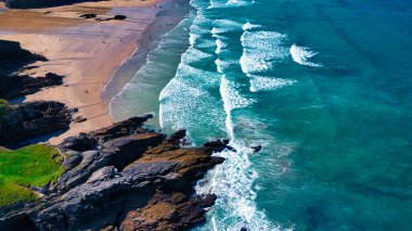 Turkuaz suları ve kıyıya vuran nazik dalgalarla dolu güzel bir sahil manzarası. Kumlu plaj, güneşten zevk alan insanlarla doludur. Kayalık oluşumlar ise manzaraya doku katar..